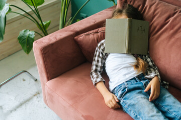 High-angle view of tired sleepy child kid girl lying on soft couch with paper book on head at home....