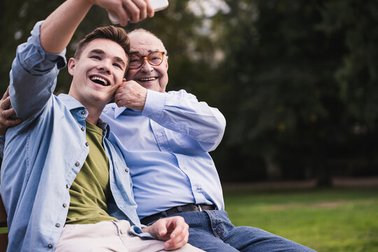 Senior man and grandson sitting together on a park bench taking selfie with smartphone
