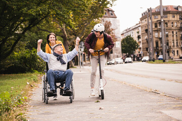 Senior man in wheelchair having fun with his adult grandchildren outdoors - obrazy, fototapety, plakaty