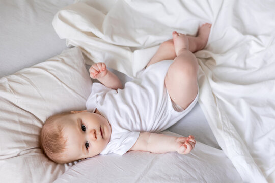 baby in a white bodysuit is lying on his back in a bed made with white bed linen. children's sleep and rest. concept of a happy childhood and motherhood. lifestyle, space for text. High quality photo