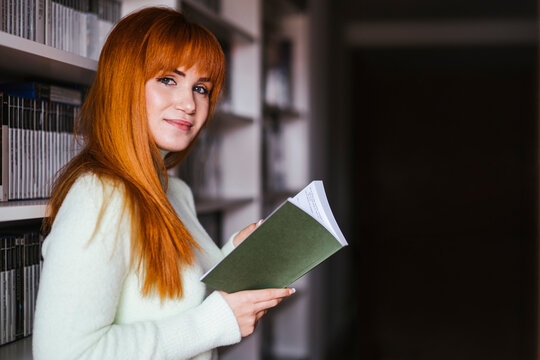 Smiling beautiful woman reading book against shelf at library