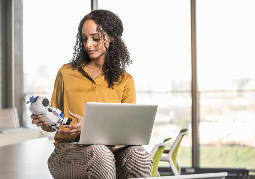 Young businesswoman sitting on desk in office with laptop and robot