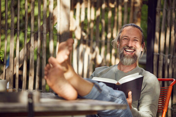 Cheerful bearded man reading book while relaxing on chair in yard