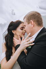 Stylish, fashionable groom in a black suit and a beautiful brunette bride in a white dress hug, kiss, standing under a long veil. Wedding portrait of lovers, smiling newlyweds.