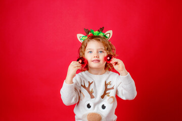 a little girl in deer horns holds Christmas tree toys on a red background, Christmas, new year