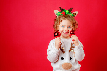 a little girl in deer horns holds Christmas tree toys on a red background, Christmas, new year
