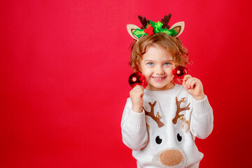a little girl in deer horns holds Christmas tree toys on a red background, Christmas, new year