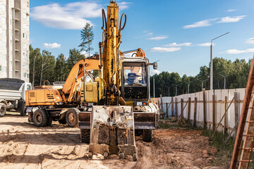 Fototapeta premium A heavy crawler excavator with a large bucket is getting ready for work in front of a panel house under construction .. Heavy construction equipment for earthworks. Quarry excavator.