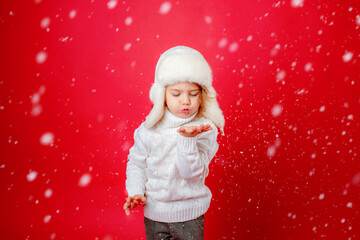 little girl in winter hat blowing on snow red background, new year.