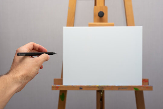 Empty white canvas stands on wooden easel on gray wall background and hand with black pencil over it  horizontal photo