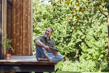 Smiling man with digital tablet looking away while sitting outside tiny house