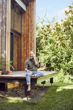 Bearded mature man talking over mobile phone while sitting outside tiny house