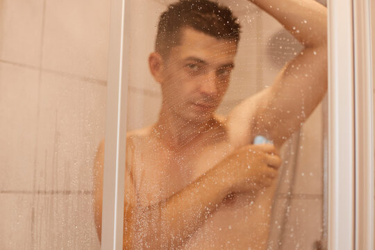 Indoor shot of attractive brunette young adult man washing his armpit with soap, looking at camera, handsome guy taking shower, refreshment after working day.