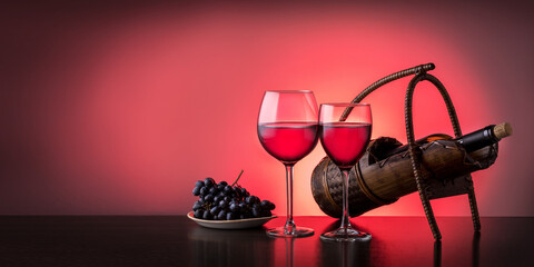 Two wine glasses with red wine and bottle of wine on the table on red background with copy space. Banner.