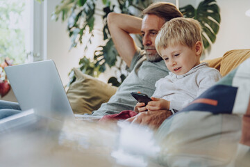 Boy using mobile phone by father with laptop in living room