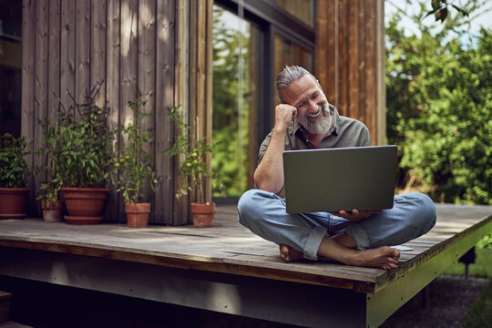 Smiling mature man using laptop while sitting against tiny house