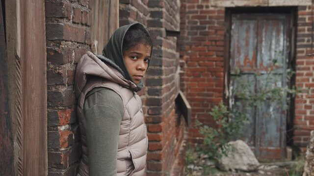 Medium portrait of abandoned 11-year-old African-American girl looking at camera standing outdoors against brick wall at poor district