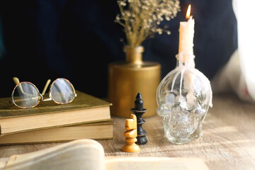 Old books, reading glasses, vintage chess pieces, lit candle and vase with gypsophila flowers. Dark academia concept. Selective focus. - obrazy, fototapety, plakaty