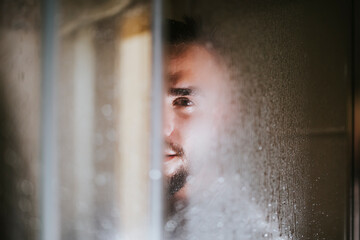 Young man looking through wet glass window from bathroom at home