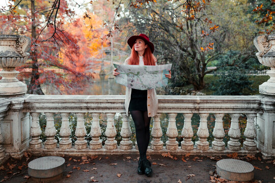 Woman wearing hat holding map while leaning on railing against lake in park