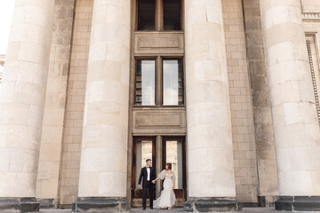 Obraz premium Young couple, man and woman, bride and groom are standing on large concrete steps among columns holding hands, looking at each other. Marriage concept of happy family life.