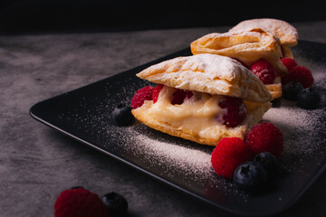 Puff pastry with cream, raspberries, blueberries and sugar powder on dark background. Delicious homemade pastry with fruit, soft cream and sugar.