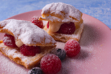 Puff pastry with cream, raspberries, blueberries and sugar powder on light blue background. Delicious homemade pastry with fruit, soft cream and sugar.