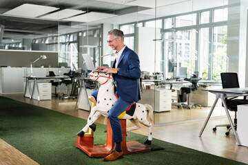 Happy male entrepreneur working on laptop while sitting on rocking horse at work place