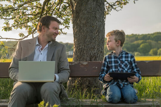 Smiling man looking at son sitting with laptop and digital tablet on bench