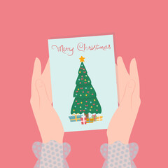 Women's hands hold a card with a Christmas tree and gifts, and the inscription Merry Christmas.