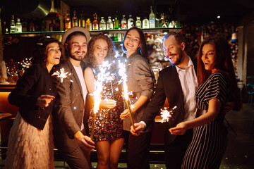 Glowing sparkles in hands. Young  friends celebrate new year and holding bengal lights and glasses with drink. Winter holiday, youth, party, lifestyle concept.