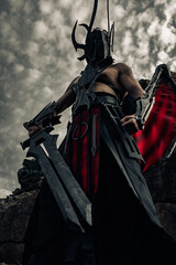 Daemon of death with wings stands with black sword among rocks against sky.