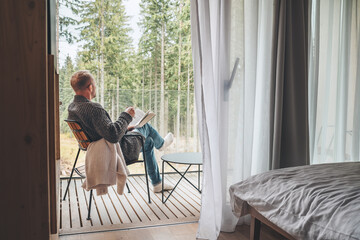 Alone man sitting in chair on country house balcony and enjoying forest view, tea cup, fresh air and bestseller novel thick book. Cozy bedroom interior back view. Reading or education concept image