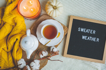 Autumn flatlay with a knitted orange sweater, a cup of hot tea, a white teapot, a burning candle, a pumpkin, a branch of cotton, letter board. Cozy.