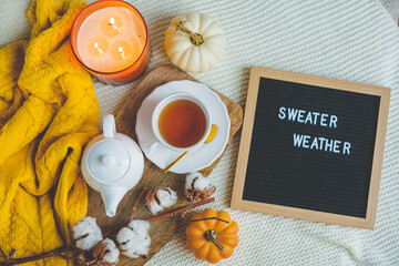 Autumn flatlay with a knitted orange sweater, a cup of hot tea, a white teapot, a burning candle, a pumpkin, a branch of cotton, letter board. Cozy.