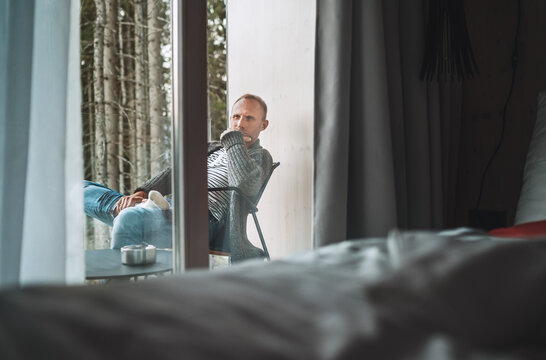 A middle-aged man dressed in open warm cardigan and jeans sitting on a forest house balcony and enjoying the fresh autumnal cold air. Everyday lifestyle concept from the room inside view image.