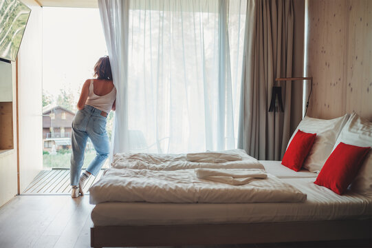 A woman dressed in blue jeans standing on forest house balcony and enjoying fresh air with nature view. Inside the Scandinavian interior design room with a king-size bed. Living in wild concept image.