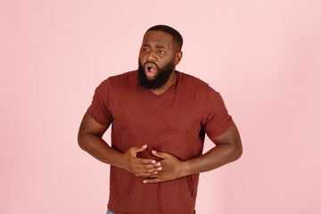 Young African-American man patient in t-shirt suffers from stomach ache standing on light pink background in studio closeup