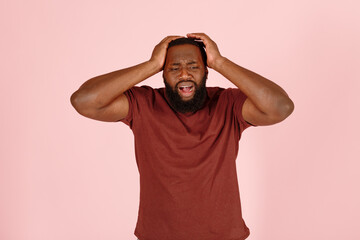 Bearded African-American man patient in t-shirt suffers from severe headache standing on pink background in studio closeup