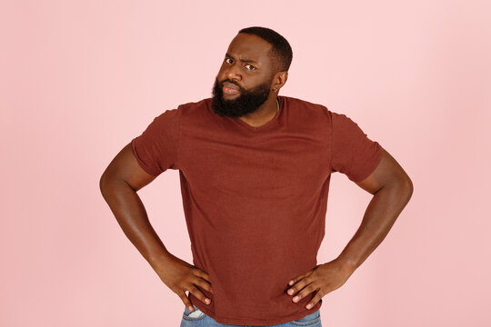 Serious bearded black man in brown t-shirt looks indignantly to camera on pink background in studio closeup