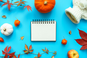 Ein leeres Notizbuch, eine Wärmflache, orange Kürbisse und Herbst Blätter auf einem blauen Hintergrund. Flat lay.