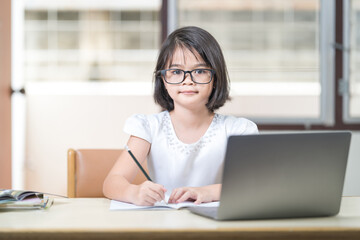 Fototapeta premium Asian children girl student with eyeglasses studying online, doing homework on laptop at home. Education Concept Stock Photo