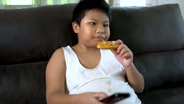 Overweight fat Asian boy sitting on sofa and eating unhealthy pizza.
