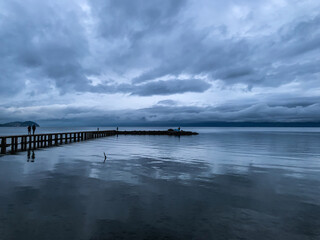Fototapeta premium Long pier at the lake, twilight, dark blue and gray sky and lake, mountains background