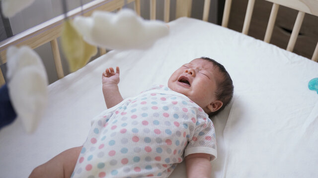 high angle shot upset newborn infant lying alone in the baby bed is crying and screaming loudly in the home nursery room.