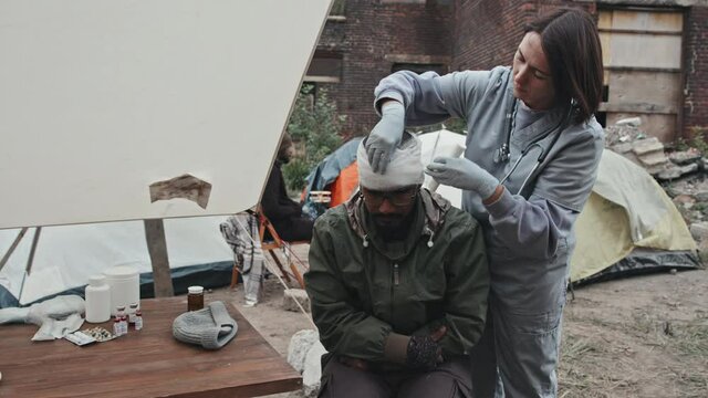 Medium shot of female medical worker bandaging head of Mixed-race man while giving first aid to refugees at tent city
