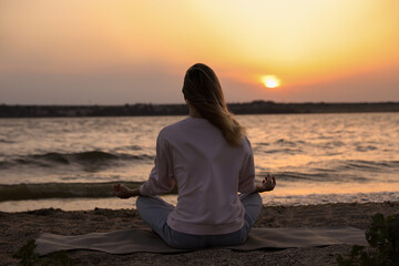 Fototapeta premium Young woman meditating on beach near river at sunset, back view