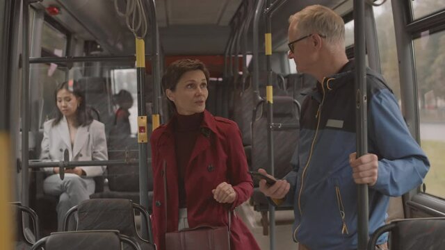 Medium of short-haired Caucasian woman wearing red trench coat holding handrail, then starting asking questions to senior male stranger, who using smartphone on bus
