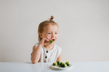 Cute funny little toddler girl eating broccoli at the table, white walls. Childhood, healthy food...