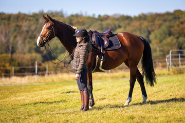 Horse with rider in full view from the side, girl stands next to the horse in a meadow, both look...